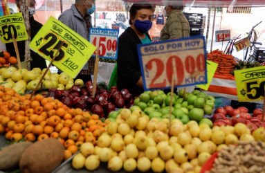 Varias personas caminado frente a un puesto de mercado popular mexicano y los letreros con precios de frutas. 