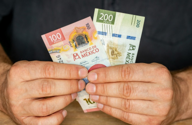 Dos manos sostienen un billete de 100 y otro de 200 pesos, al estar recargados sobre una mesa. 