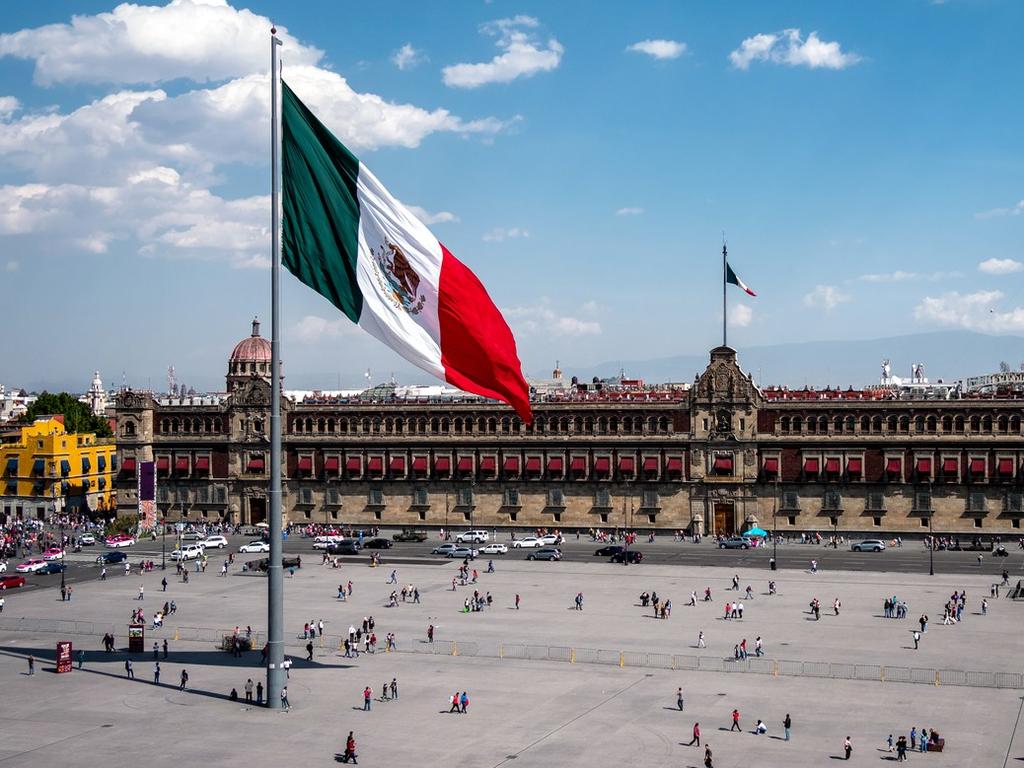Bandera de México en la plancha del Zócalo capitalino con personas caminando. 
