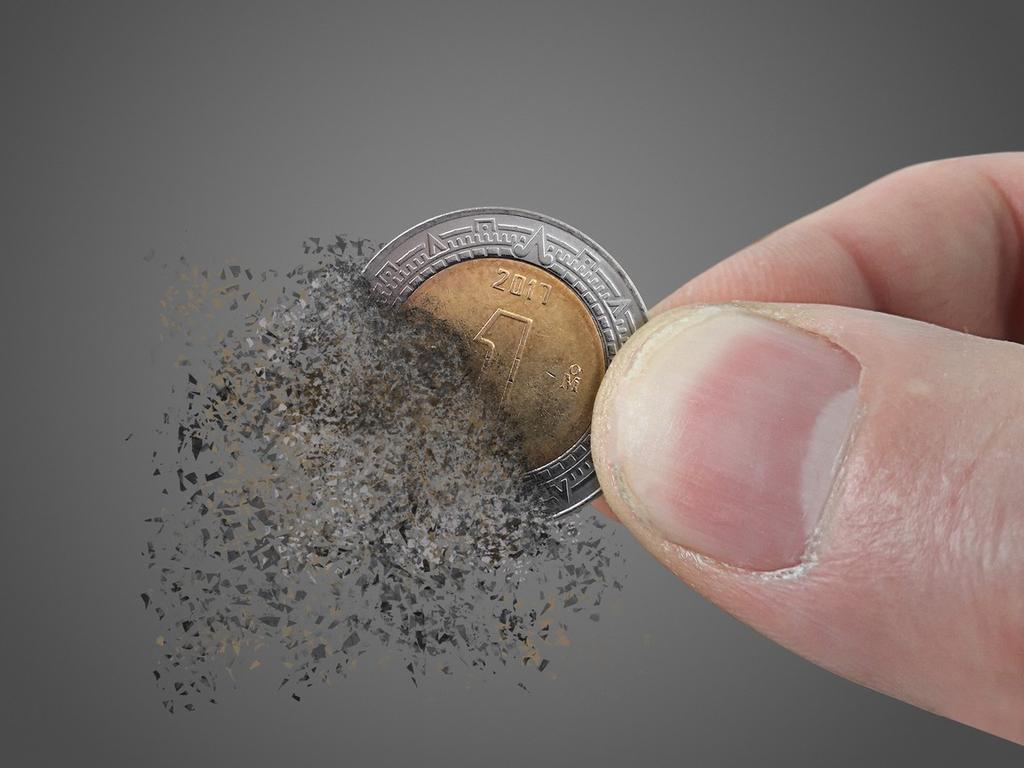 Dedos sostienen una moneda de un peso mexicano que se despedaza, en un fondo color gris. 