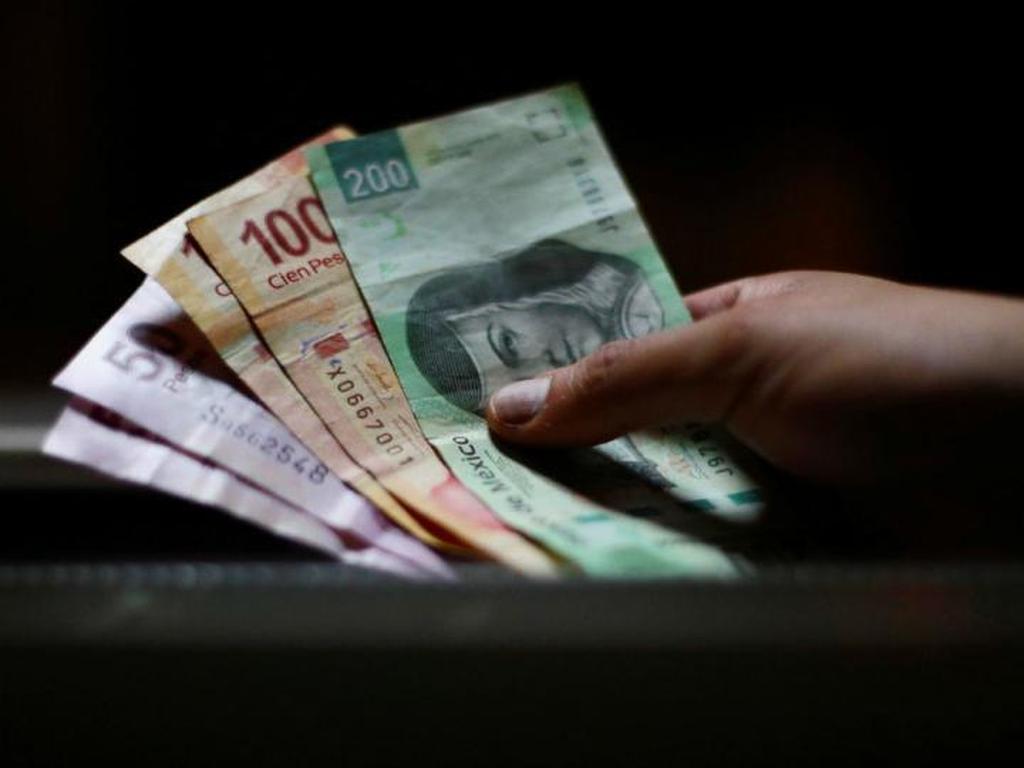 Una mano sostiene billetes de 200, 100 y 50 pesos mexicanos en un fondo oscuro. 