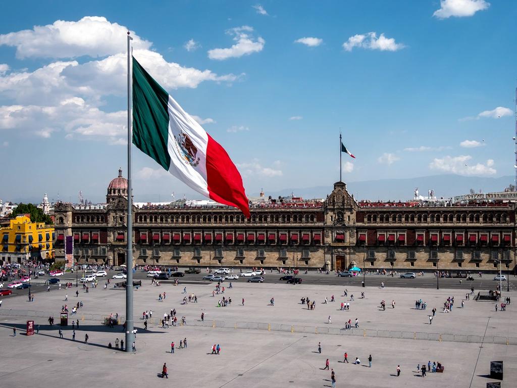 La bandera de México en el Zócalo con personas caminando y el Palacio Nacional. 
