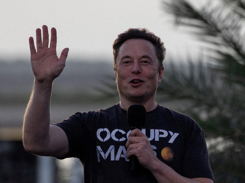 El empresario Elon Musk sostiene un micrófono para hablar y alza la otra mano. 