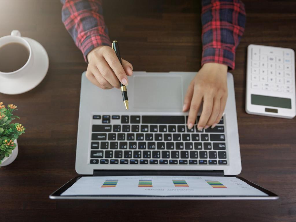 manos de mujer en una computadora sobre mesa cafe, con calculadora y taza de cafe a los lados