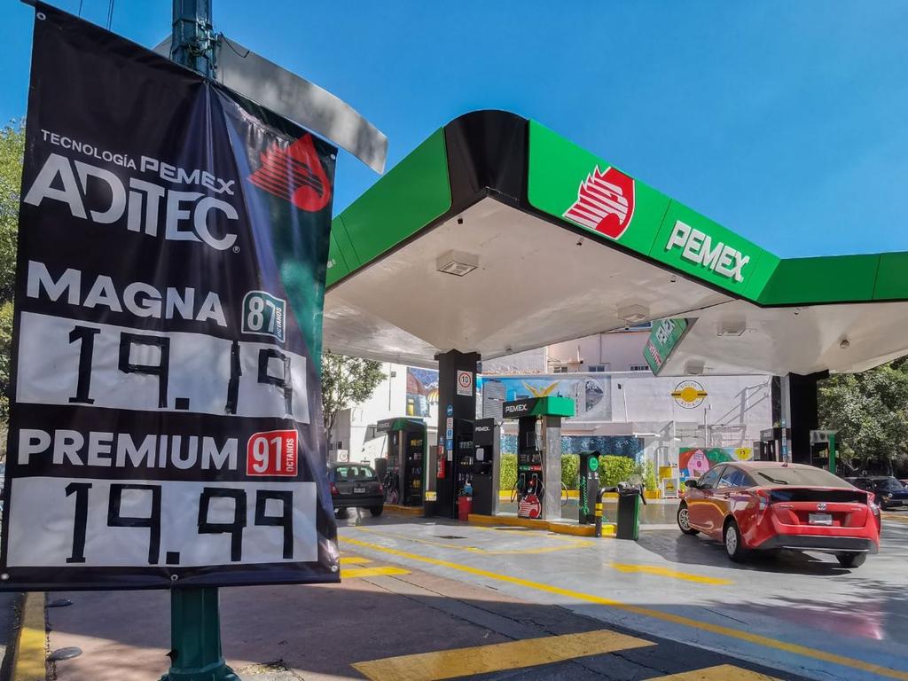 Un auto color rojo llega a una gasolinera de la marca Pemex y un letrero con los precios. 