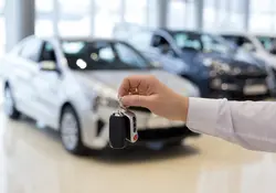 Una mano sostiene las llaves de un automóvil y de fondo se observan varios estacionados. 