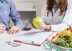Doctor con manzana en mano sobre una mesa una tabla con papeles, lentes y ensalada