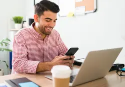 Un hombre sonríe al utilizar su teléfono celular al estar sentado frente a una computadora. 