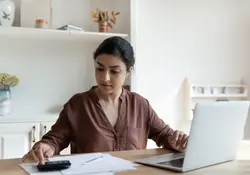 mujer enfocada haciendo cuentas con calculadora y computadora sobre la mesa 