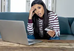 joven mujer conflictuada con tarjeta de credito en mano y computadora de frente en una sala