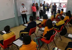 Salón de clases con estudiantes sentados en las butacas y autoridades paradas al frente con un pizarrón blanco. 