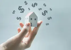Modelo de casa en mano de una persona con icono de dinero alrededor sobre fondo gris 