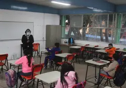 Alumnos sentados en un salón de clases y una maestra parada al frente. 