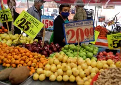 Personas caminan en un mercado popular con la venta de frutas y verduras, hay letreros con los precios de los alimentos. 