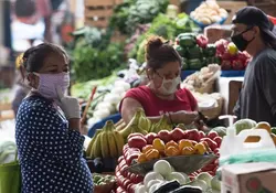 Dos señoras realizan compras de verduras y frutas en un mercado popular, una persona pasa caminando al fondo. 
