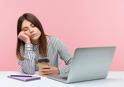 Una mujer duerme mientras trabaja frente a una laptop y con su café en la mano. 