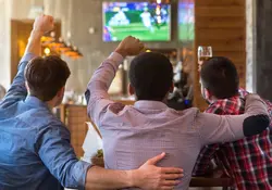 Tres hombres de espaldas celebran con las manos al observar un partido de futbol desde un bar. 