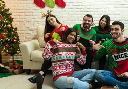 Tres mujeres y dos hombres visten ugly sweater al posar frente a la cámara, se encuentran sentados en una sala con un árbol de Navidad al costado. 