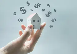 Una mano sostienen pequeña figura de una casa y encima tiene varios signos de dinero flotando. 