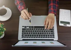 manos de mujer en una computadora sobre mesa cafe, con calculadora y taza de cafe a los lados