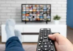 La mano de una persona sostiene un control remoto dirigido hacia una smart tv, específicamente en la sala de una casa. 