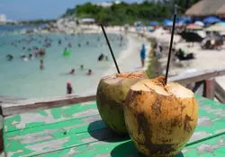 do cocos con popotes sobre una mesa verde con fondo borroso de la playa y personas