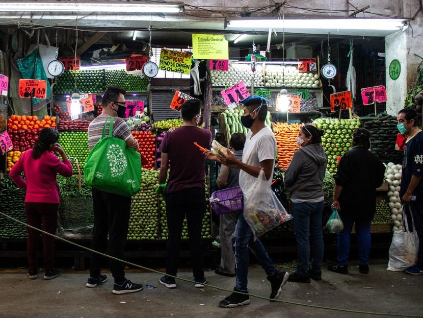 Varias personas paradas frente a un puesto de verduras en un mercado. 