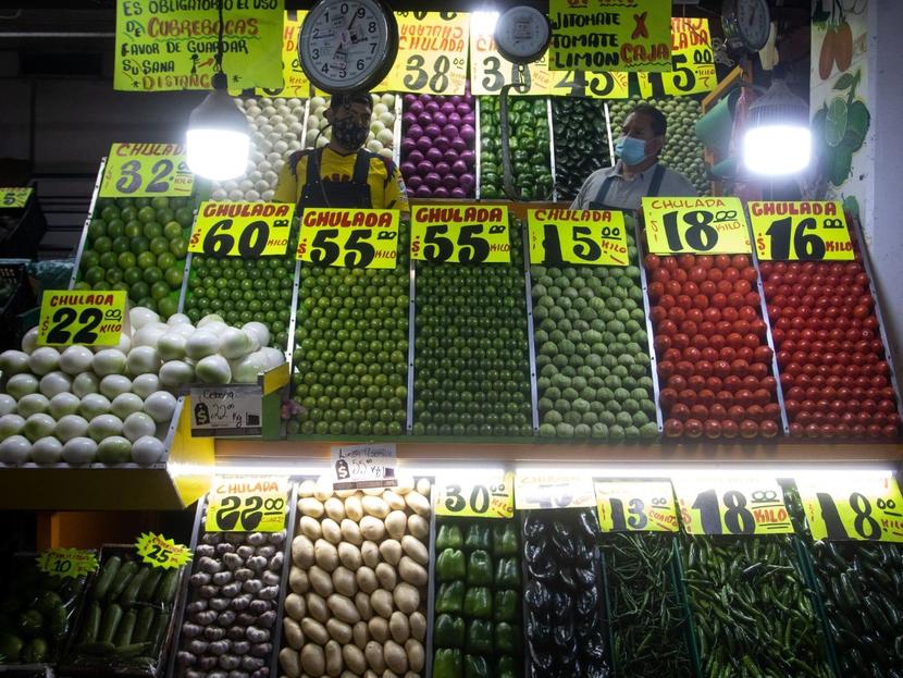 Dos personas atienden un puesto de verduras en un mercado y hay letreros con los precios de cada producto. 