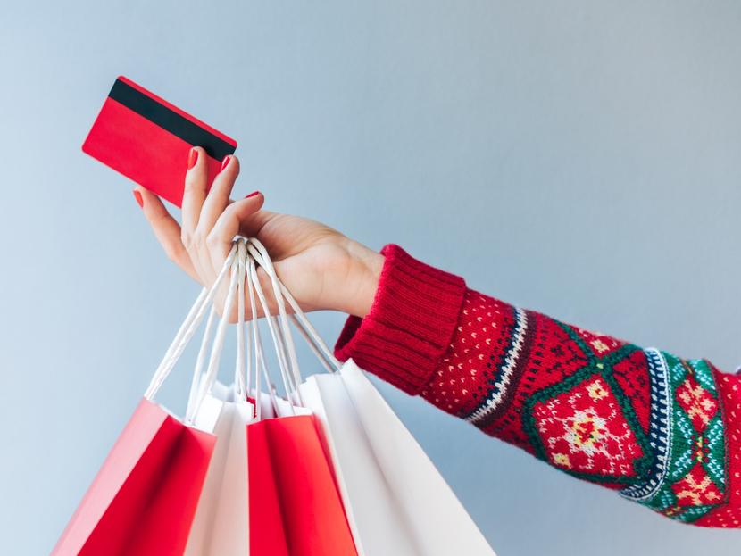 Un brazo con suéter de navidad sostiene varias bolsas de tiendas y una tarjeta bancaria color rojo. 