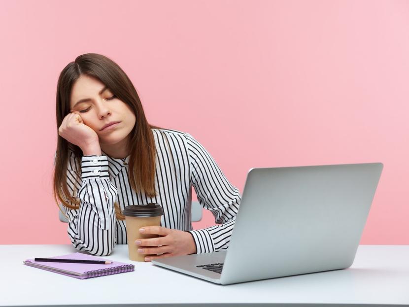 Una mujer duerme mientras trabaja frente a una laptop y con su café en la mano. 