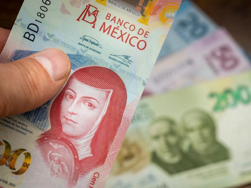 Una mano sostiene un billete de 100 pesos mexicanos, al fondo hay billetes de 200, 50 y 20 pesos. 