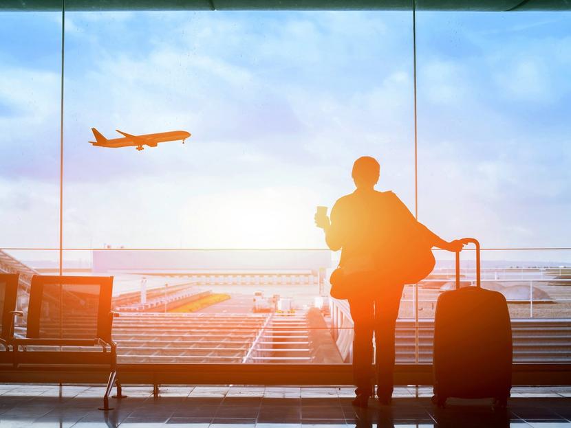 Una persona parada con su maleta y con un café en un aeropuerto, al mismo tiempo observa el despegue de un avión. 