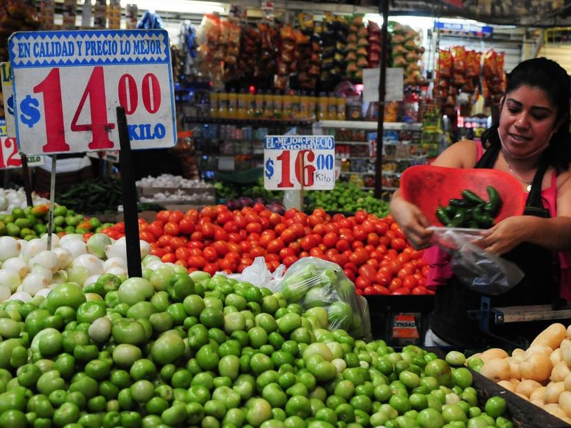 Una mujer atiene un puesto e verduras en un mercado popular, ahí hay letreros con los precios de cada producto. 