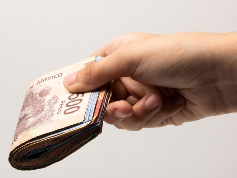 Una mano sostiene un fajo de billetes de pesos mexicanos de distintas denominaciones. 