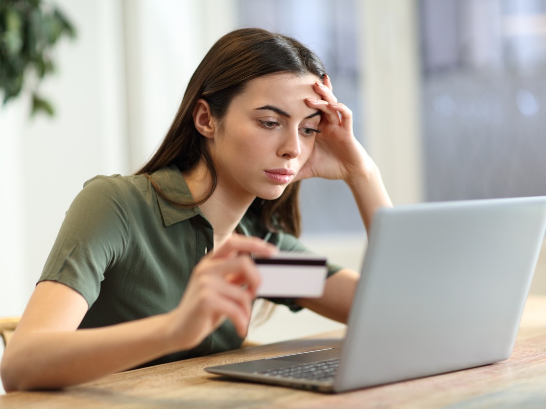 Una mujer observa preocupada una laptop y sostiene una tarjeta bancaria. 