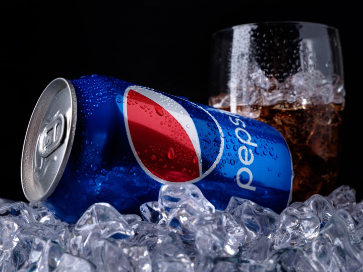 Una lata de refresco azul marca Pepsi colocada sobre varios hielos y un vaso de vidrio al costado. 