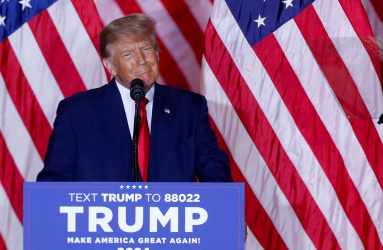 El expresidente de los Estados Unidos Donald Trump sonríe al estar parado en un escenario, frente a un micrófono  y banderas atrás. 