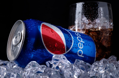 Una lata de refresco azul marca Pepsi colocada sobre varios hielos y un vaso de vidrio al costado. 