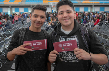 Dos jóvenes sonríenal sostener un sobre rojo de la Beca Benito Juárez, hay muchas personas y sillas al fondo. 