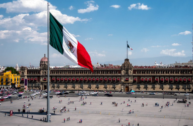 Plancha del Zócalo con la bandera de México y muchas personas caminando alrededor. 