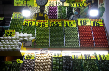 Dos personas atienden un puesto de venta de verduras y hay letreros de los precios por kilo. 